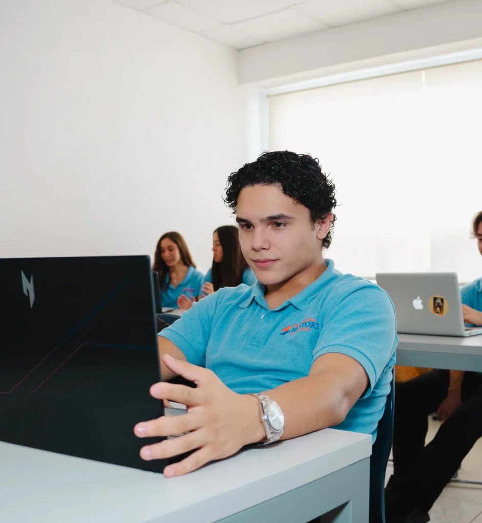 Estudiante de Newport High School con polo azul trabajando en su laptop en el salón de clases, con compañeros al fondo